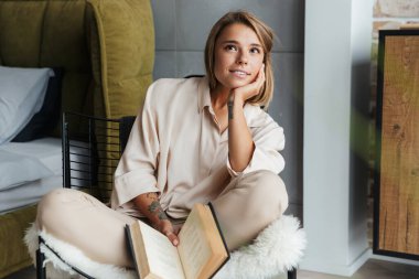 Oturma odasındaki koltukta otururken şık pijamalarıyla kitap okuyan beyaz bir kadının resmi.