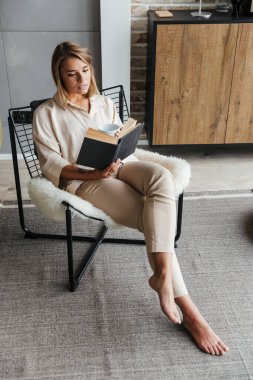 Oturma odasındaki koltukta oturmuş çay içip kitap okuyan genç bir kadın resmi.