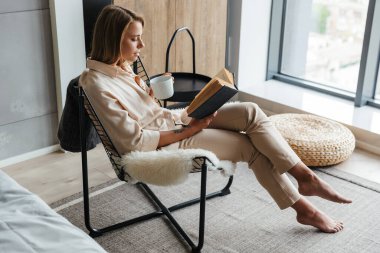 Oturma odasındaki koltukta oturmuş çay içip kitap okuyan genç bir kadın resmi.