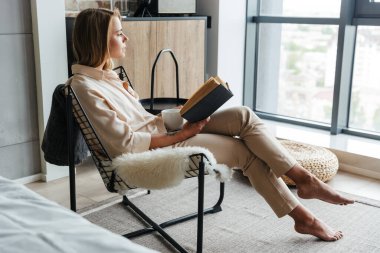 Oturma odasındaki koltukta oturmuş çay içip kitap okuyan genç bir kadın resmi.