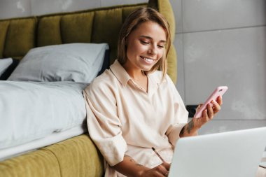 Pijamalı neşeli bir kadının yatak odasında yerde otururken dizüstü bilgisayar ve akıllı telefon kullanması.