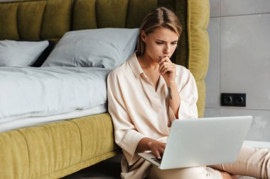Düşüncelere dalmış, pijamalı, laptopla çalışan, yatak odasında yerde oturan güzel bir kadın.