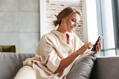 Pijamalı, gülümseyen ve koltukta otururken cep telefonu kullanan mutlu bir kadın resmi.