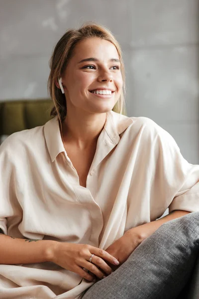 パジャマ姿の幸せな女性の笑顔とリビングでソファに座っている間の無線イヤフォンの使用のイメージ — ストック写真