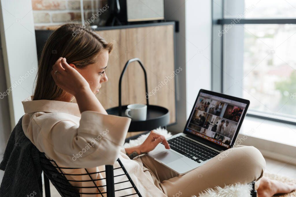 Image of focused blonde nice woman in stylish pajamas working with laptop while sitting on armchair at living room