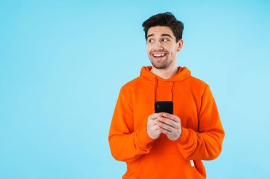 Kapüşonlu genç bir adamın portresi mavi arka planda izole bir şekilde duruyor, cep telefonunu tutuyor, başka tarafa bakıyor.