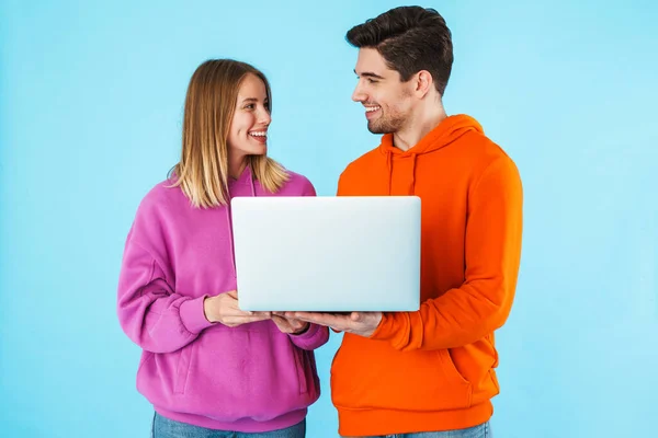 一对笑容满面的年轻夫妇的画像 他们穿着帽衫 孤零零地站在蓝色的背景下 用笔记本电脑 — 图库照片