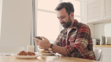 Yakışıklı, genç, pozitif sakallı bir adam evde evde kahvaltı ederken cep telefonu ve kahve içiyor.