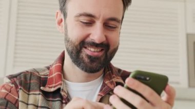 Mutfakta cep telefonuyla gülümseyen yakışıklı, sakallı bir adam.