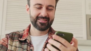 Evde kahve içerken cep telefonu kullanan yakışıklı, konsantre sakallı bir adam.