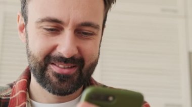 Yakışıklı, konsantre ve pozitif sakallı bir adam. Evdeki mutfakta cep telefonu kullanıyor.