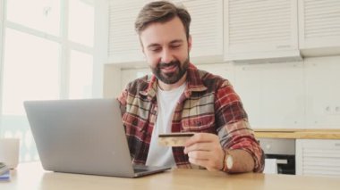 Yakışıklı, mutlu, sakallı bir adam elinde kredi kartı tutarken dizüstü bilgisayar kullanıyor.