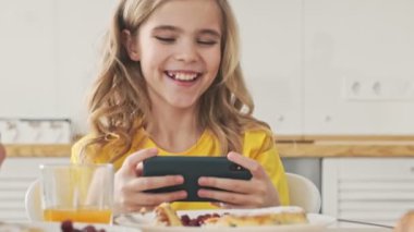 Neşeli, mutlu bir genç kız akıllı telefonuyla dijital oyunlar oynuyor. Sabahleyin mutfakta kahvaltıda onu yatay olarak tutuyor.