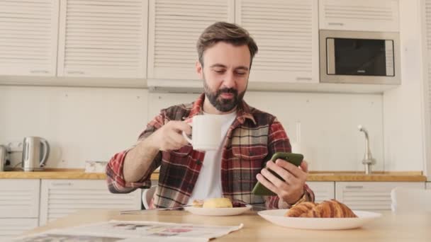 在家里厨房边吃早饭边用手机喝茶或喝咖啡的英俊而快乐的小胡子男人 — 图库视频影像