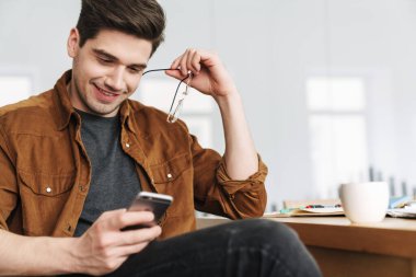 Ofiste kahve içerken gülümseyen ve cep telefonu kullanan yakışıklı bir genç resmi