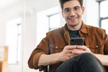 Ofiste çalışırken gülümseyen ve cep telefonu kullanan yakışıklı, neşeli bir genç resmi