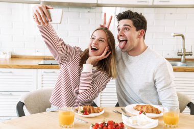 Evdeki rahat mutfakta kahvaltı ederken akıllı telefondan selfie çeken genç çiftin portresi.