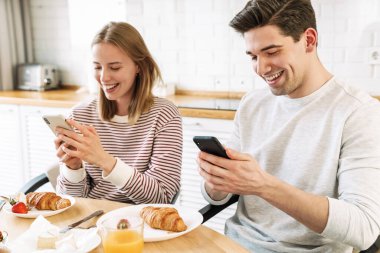 Evde rahat bir mutfakta kahvaltı ederken akıllı telefon kullanan genç bir çiftin portresi.