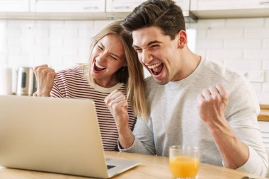 Evde rahat bir mutfakta kahvaltı ederken dizüstü bilgisayar kullanan ve kazanan jestler yapan heyecanlı genç çiftin portresi.