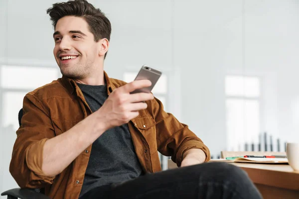 Imagen Joven Guapo Alegre Sonriendo Usando Teléfono Celular Mientras Bebe — Foto de Stock