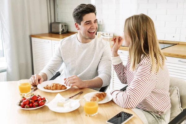 Portrait of young happy couple eating together at table while having breakfast in cozy kitchen at home