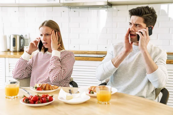 Portrait of young confused couple talking on smartphones while having breakfast in cozy kitchen at home