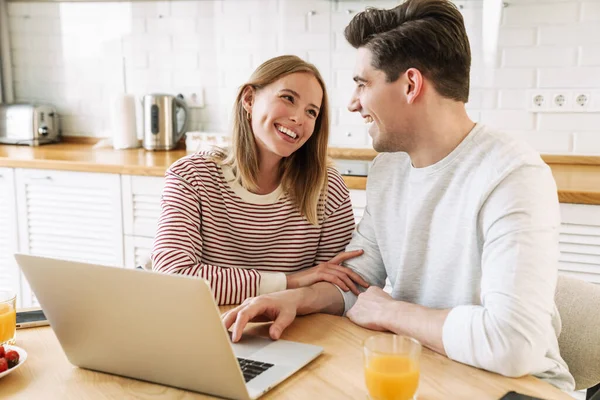 Portrait of young laughing couple using laptop and talking while having breakfast in cozy kitchen at home