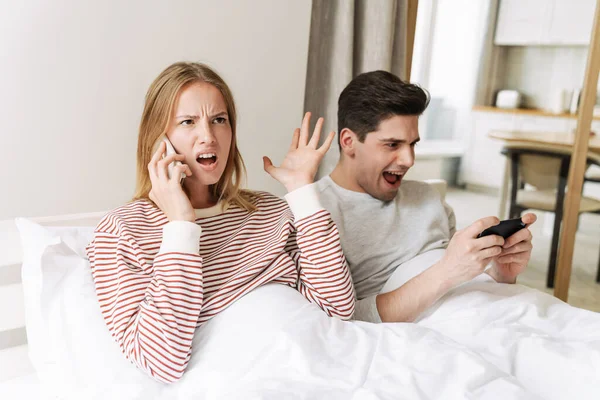 Portrait of beautiful emotional couple using mobile phones while lying in bed at home