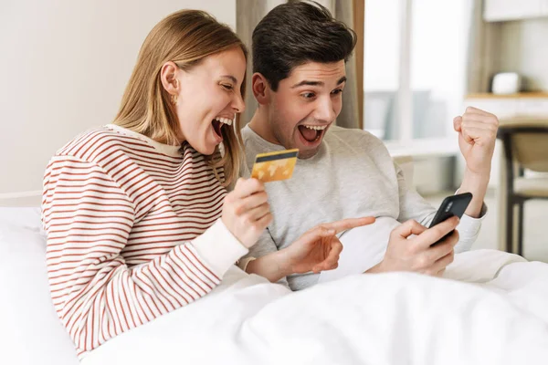 Portrait of beautiful excited couple holding smartphone and credit card while making winner gesture in bed
