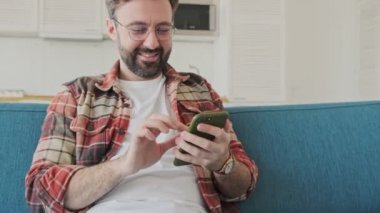 Yakışıklı, gülümseyen, mutlu, sakallı bir adam evde koltukta oturuyor ve cep telefonu kullanıyor.