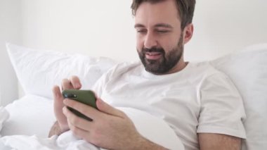 Yakışıklı, konsantre olmuş sakallı adam evde, akıllı telefon kullanırken yatakta yatıyor.