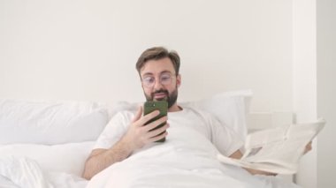 Yakışıklı genç sakallı adam evde yatıp gazete okurken eline bir cep telefonu alıp yatağın üzerine gazete koyuyor.
