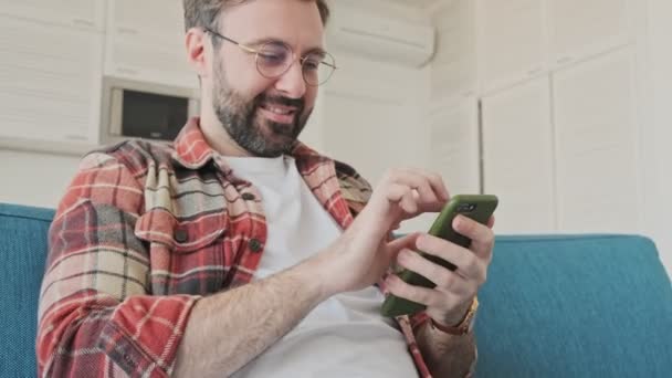 携帯電話を使用している間にソファに座っている家の中で楽しい髭の男を屋内に集中ハンサムな若い — ストック動画