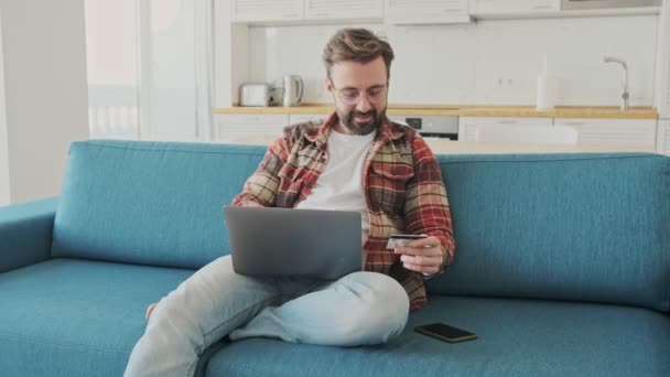 ノートパソコンを使いクレジットカードを持ちながらソファに座っている家の中にいるハンサムな若い髭の男 — ストック動画