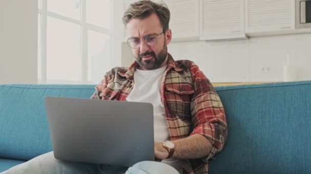 Evdeki Kanepede Dizüstü Bilgisayar Kullanarak Oturan Yakışıklı Yorgun Sakallı Adam — Stok video
