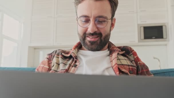ラップトップコンピュータを使用している間にソファに座っている家の中でハンサムな若い濃縮ひげそりの男 — ストック動画