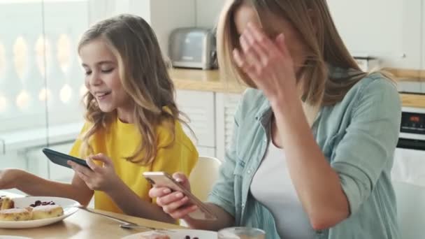 一个年轻漂亮的姑娘和她的妈妈早上在厨房里吃早饭时正在用她们的智能手机 — 图库视频影像