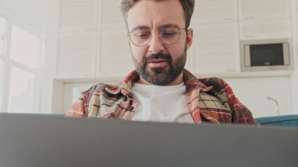 ノートパソコンを使用している間にソファに座っている家の中でハンサムな若い疲れた髭の男 — ストック動画