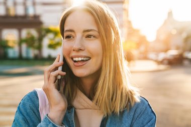 Kot ceket giyen güzel sarışın kadın şehirde yürüyor, cep telefonuyla konuşuyor.