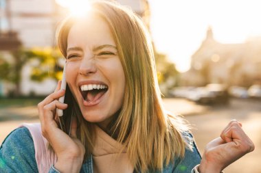 Kot ceket giyen güzel sarışın kadın şehirde yürüyor, cep telefonuyla konuşuyor, kutlama yapıyor.