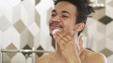 Genç, yakışıklı, pozitif Afrikalı bir adam tuvalette aynanın yanında cildine iyi bakıyor.