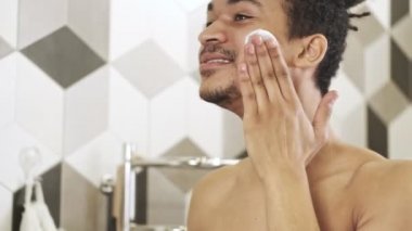 Genç, yakışıklı, mutlu, Afrikalı bir adam tuvalette aynanın yanında cildine bakıyor.