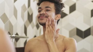 Genç, yakışıklı, iyimser Afrikalı adam banyoda aynanın yanında cildine krem sürüyor.