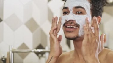 Genç, iyimser Afrikalı bir adam banyoda aynanın yanında yüzüne maske takıyor.