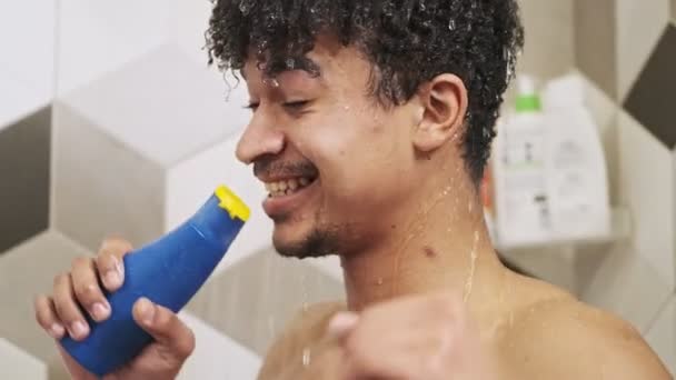 シャワージェル付きのボトルに歌うバスルームで室内でシャワーでアフリカの感情的な幸せな男 — ストック動画