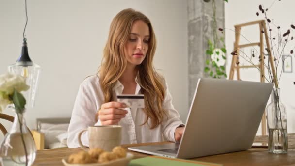 かなり素敵な若い女性は 自宅で居心地の良い部屋で朝食中にオンラインショッピングに行く彼女のクレジットカードの数を彼女のラップトップコンピュータに入力しています — ストック動画