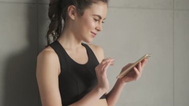 Siyah eşofmanlı, gülümseyen, pozitif bir kadın sabah gri bir stüdyoda çalışmadan önce akıllı telefonunu duvarın yanında kullanıyor.