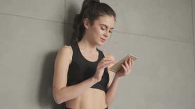 Siyah eşofmanlı güzel bir kadın sabah gri bir stüdyoda çalışmadan önce akıllı telefonunu duvarın yanında kullanıyor.