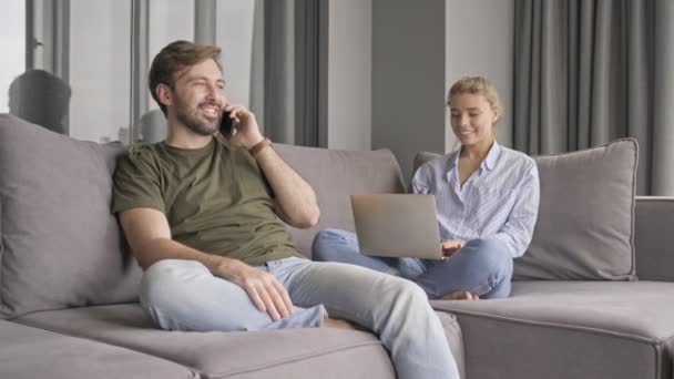 魅力的な白人の若いカップルは一緒に自宅で時間を費やしていますリビングルームのソファに座って — ストック動画