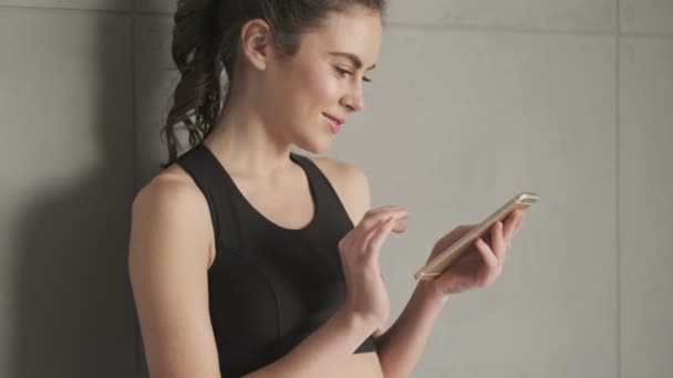 一位穿着黑色运动服 面带微笑 积极向上的年轻女子 早上在一家灰色工作室接受训练之前 正在使用她的智能手机站在墙边 — 图库视频影像